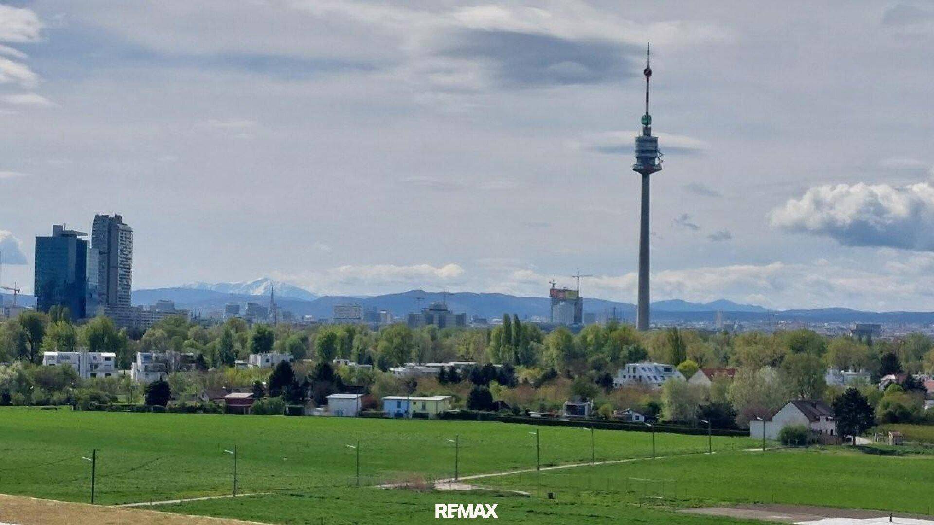 Panoramablick über die Stadt mit Donauturm und Alpen im Hintergrund unter bewölktem Himmel.