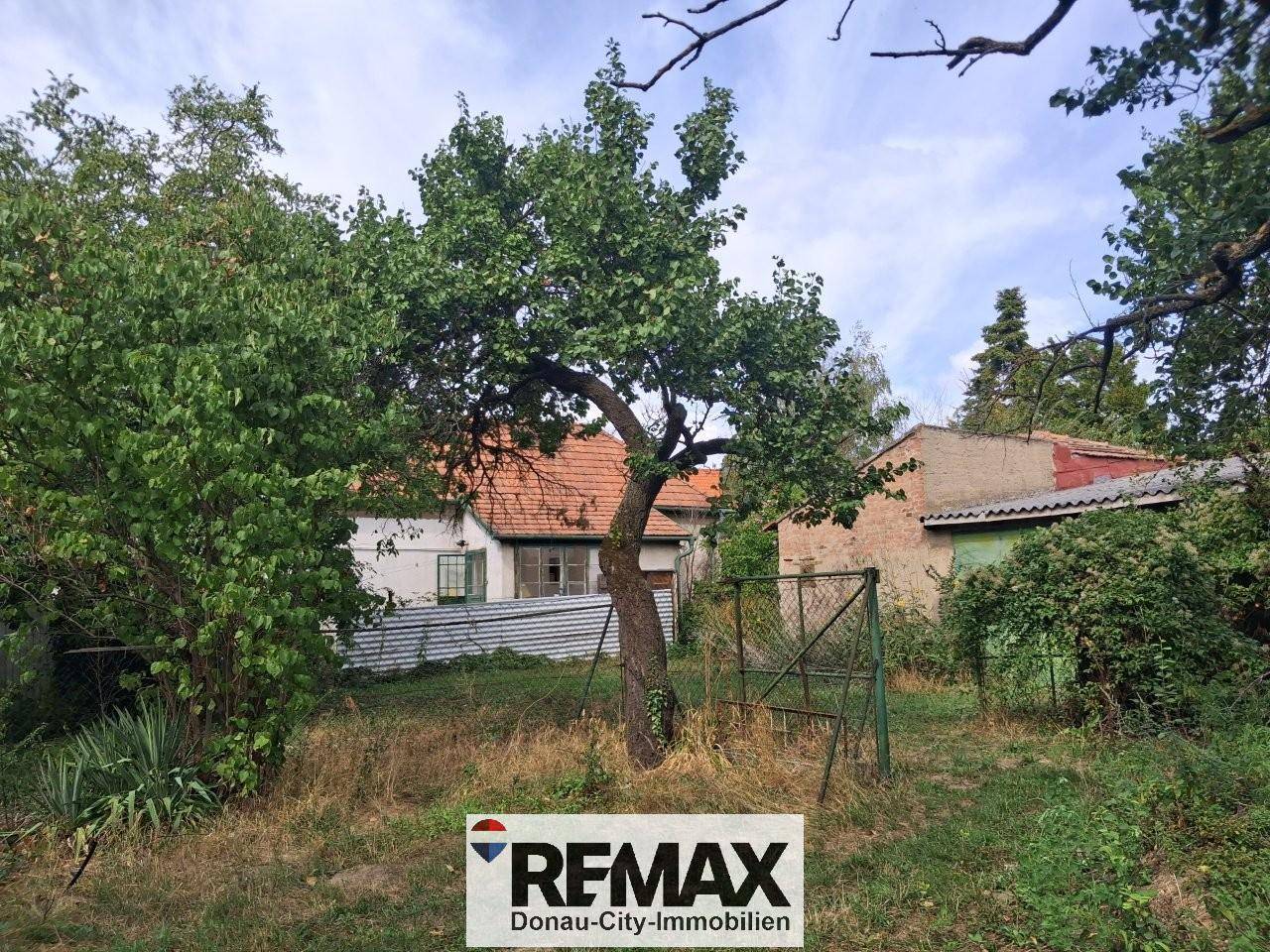 Blick auf ein älteres Haus mit rotem Ziegeldach, umgeben von dichtem Grün und Bäumen.