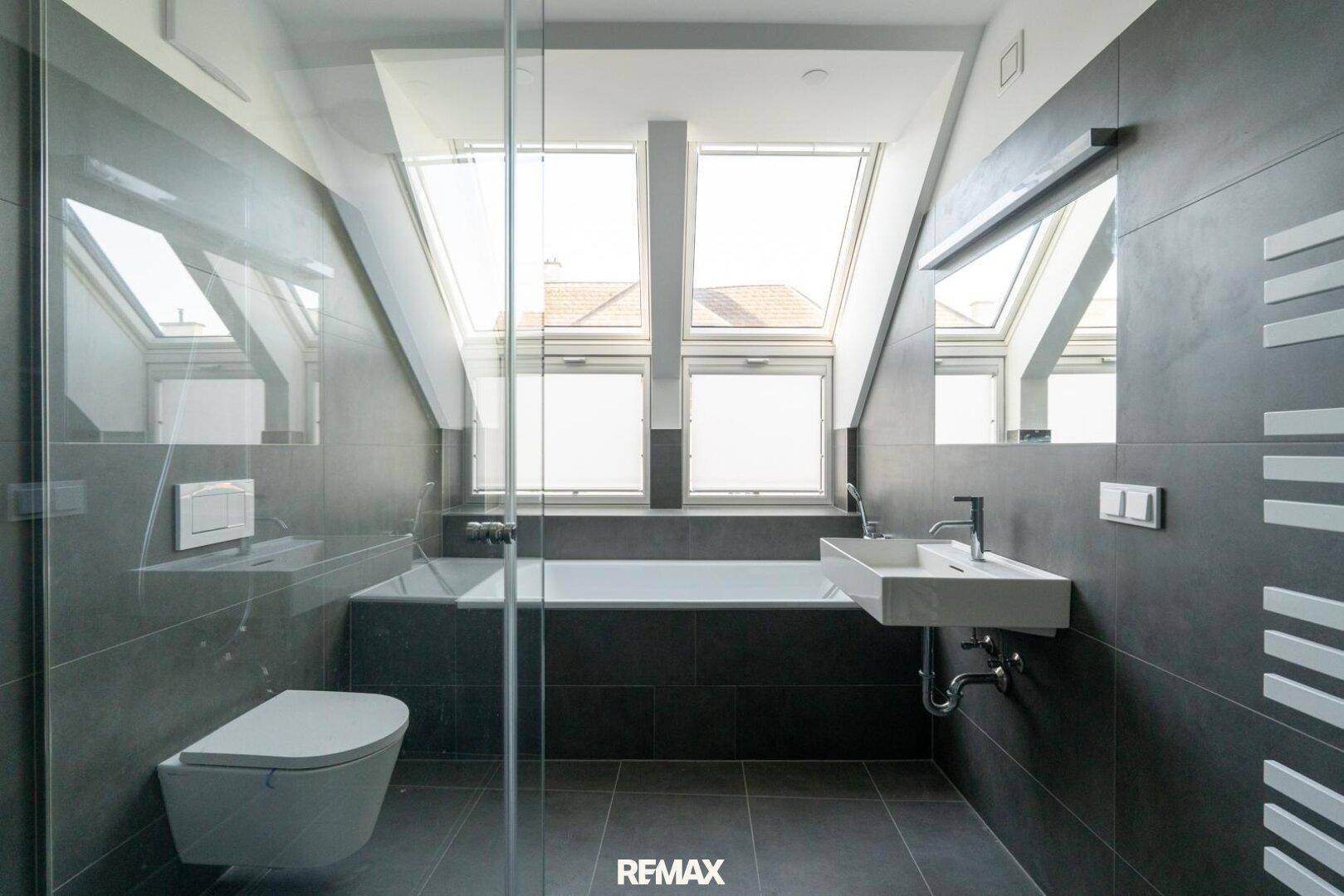 Modernes Badezimmer mit Badewanne, Dusche, Dachfenstern und grauen Fliesen.