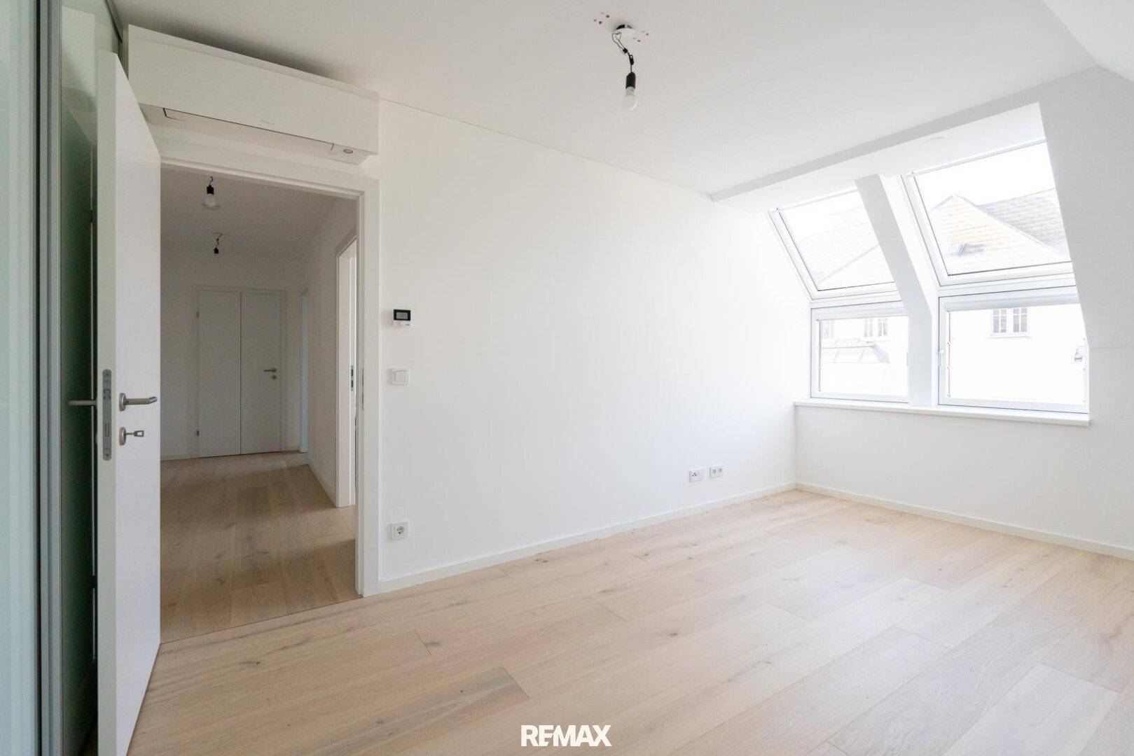 Geräumiges Schlafzimmer mit Dachschrägen, hellem Parkettboden und Zugang zu einem weiteren Raum.