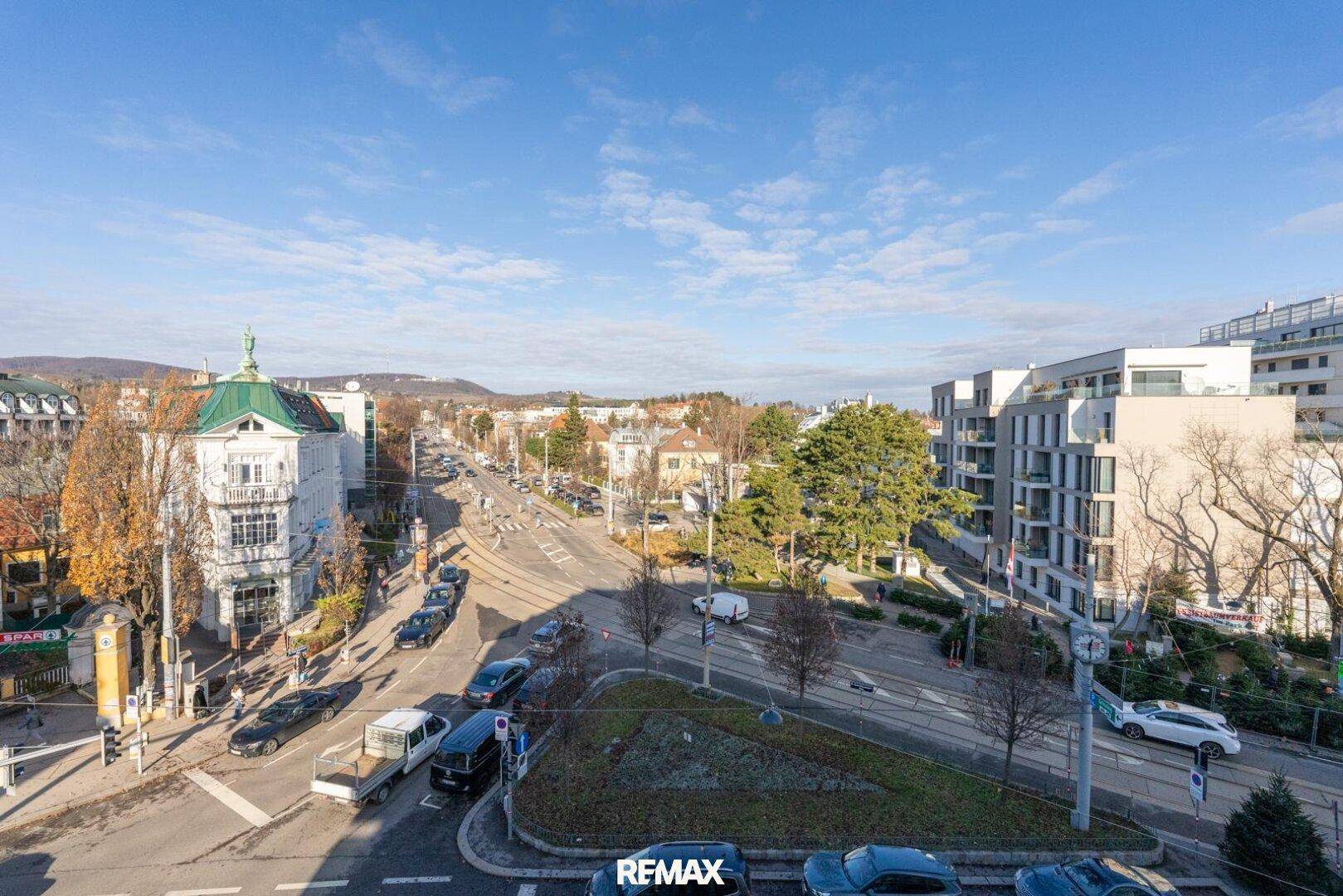 Weitläufiger Blick über eine belebte Stadtkreuzung mit viel Grün und Gebäuden im Hintergrund.