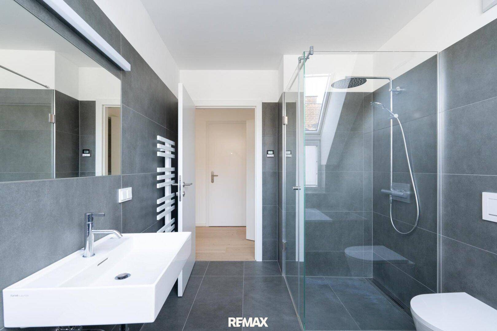 Elegantes Badezimmer mit Dusche, Waschbecken, großem Spiegel und grauen Fliesen.