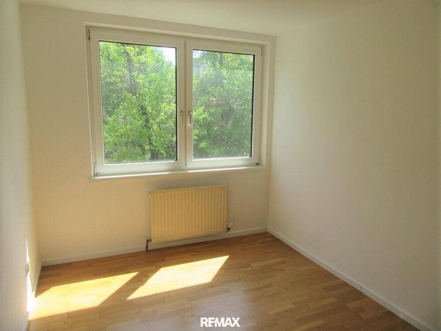 Freundliches Schlafzimmer mit Parkettboden und Fenster mit Blick auf grüne Bäume.
