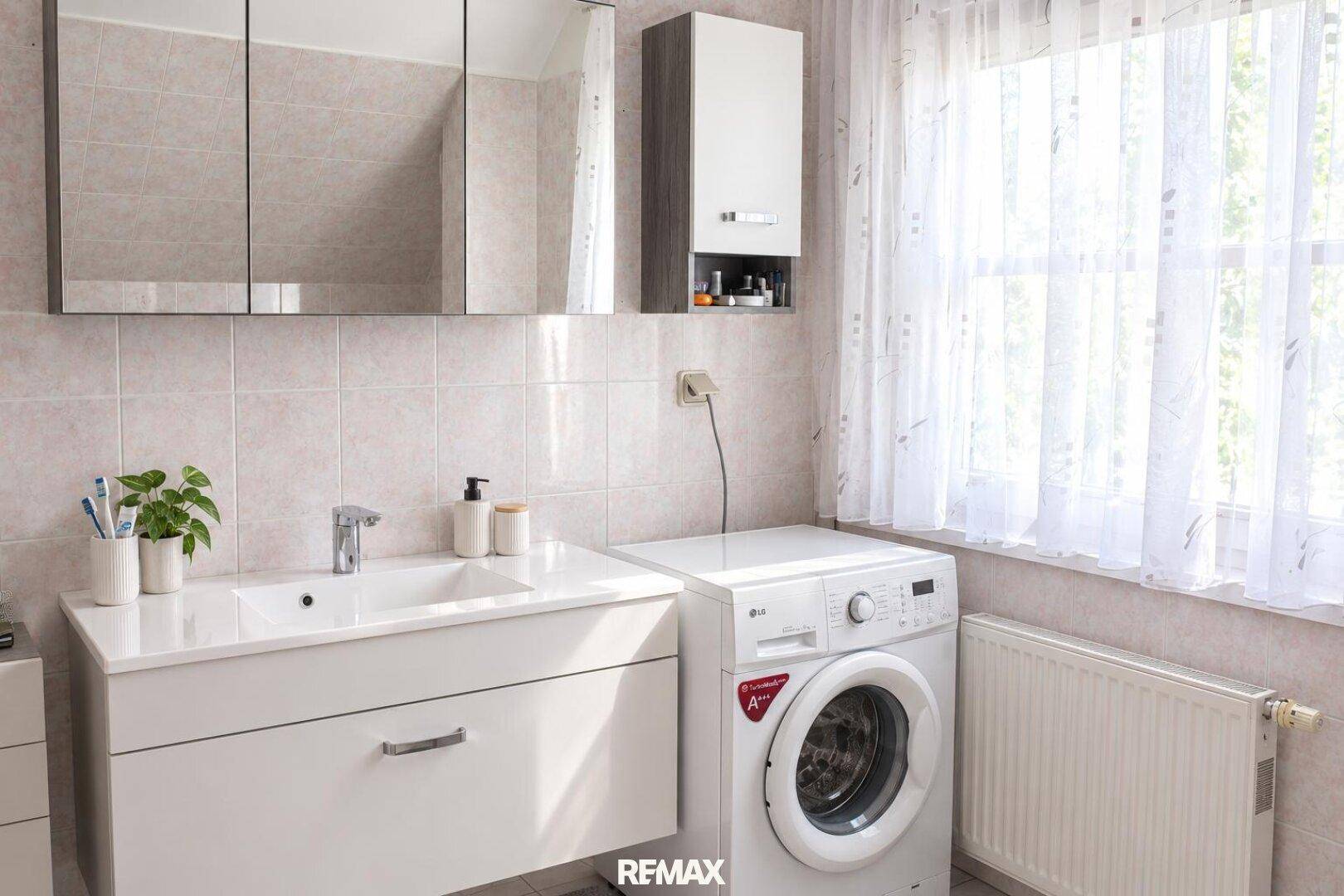 Badezimmer mit Waschmaschine, Waschtisch, Spiegelschrank und Fenster.