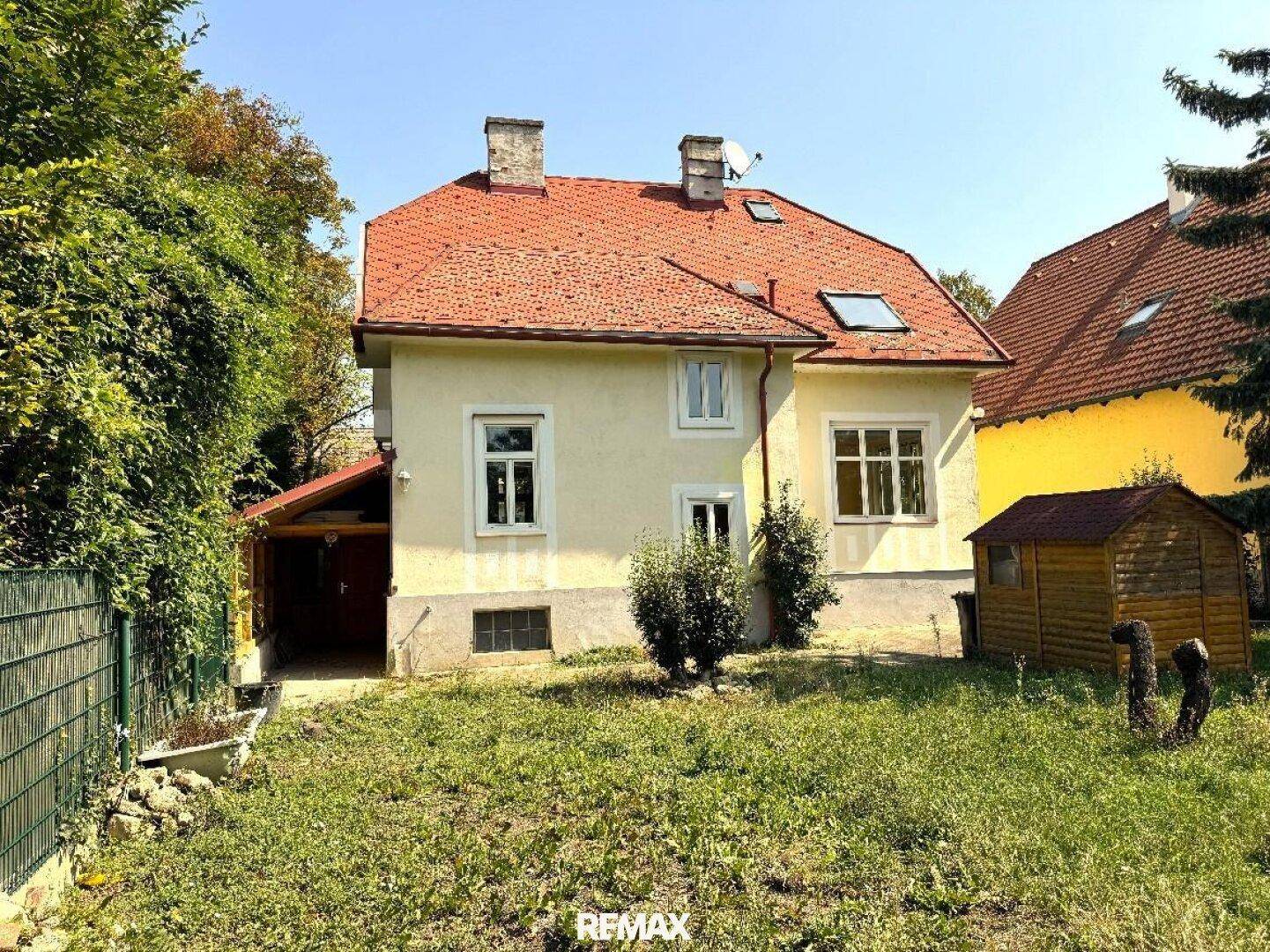 Das Haus mit gelber Fassade und rotem Dach, umgeben von einem grünen Garten und einem kleinen Holzschuppen.