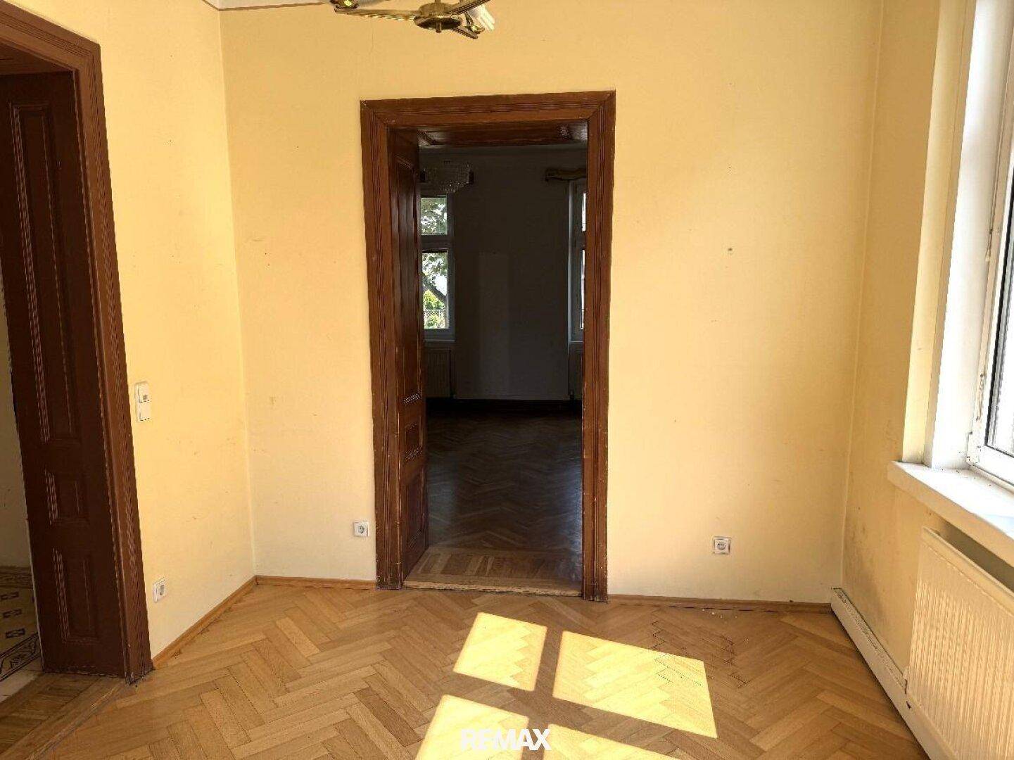 Helles Zimmer mit Fischgrätparkett und gelben Wänden, das durch eine Holztür mit einem weiteren Raum verbunden ist.