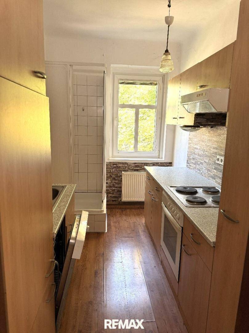 Schmale Küche mit hellen Holzschränken, integrierter Duschkabine und Fenster, praktisch und funktional.