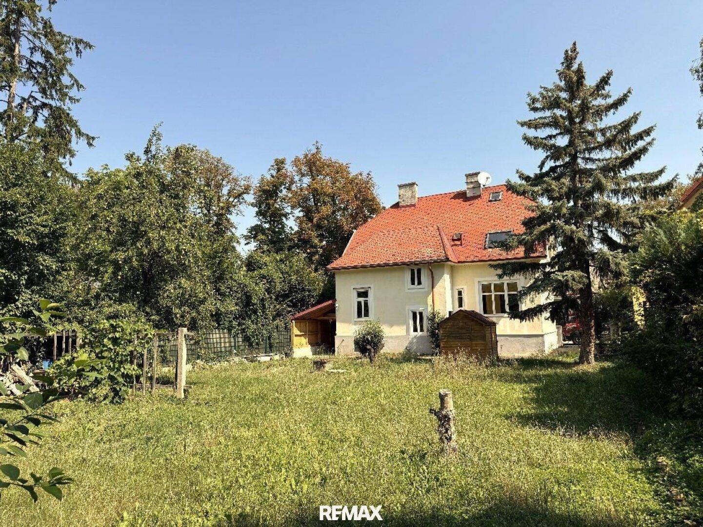 Ansicht des Hauses vom Garten aus, mit gelber Fassade, rotem Dach und üppigem Grün unter blauem Himmel.