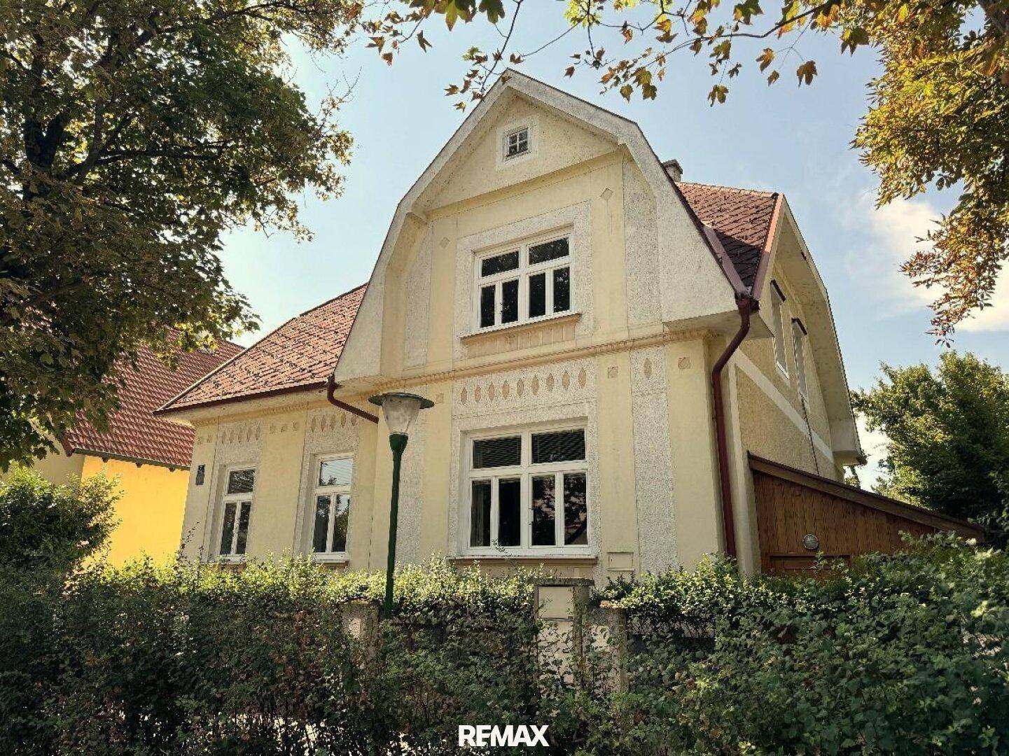 Vorderansicht des Hauses von der Straße, mit gelber Fassade, rotem Dach und einem gepflegten Zaun mit Hecke.