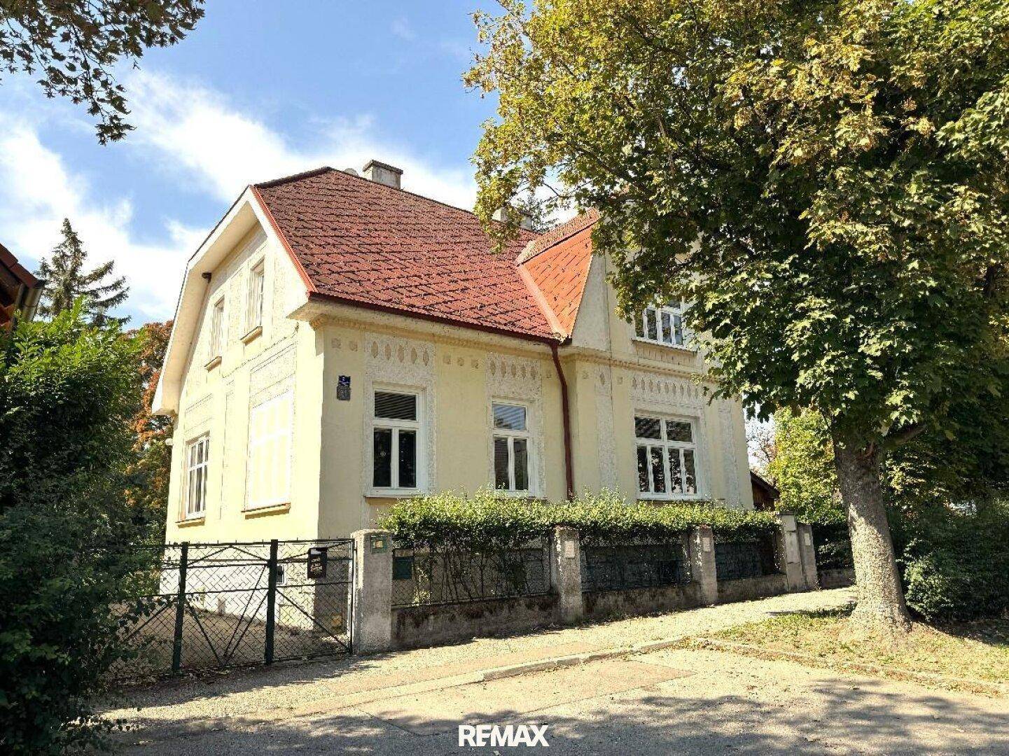 Vorderansicht des Hauses von der Straße, mit gelber Fassade, rotem Dach und einem gepflegten Zaun mit Hecke.