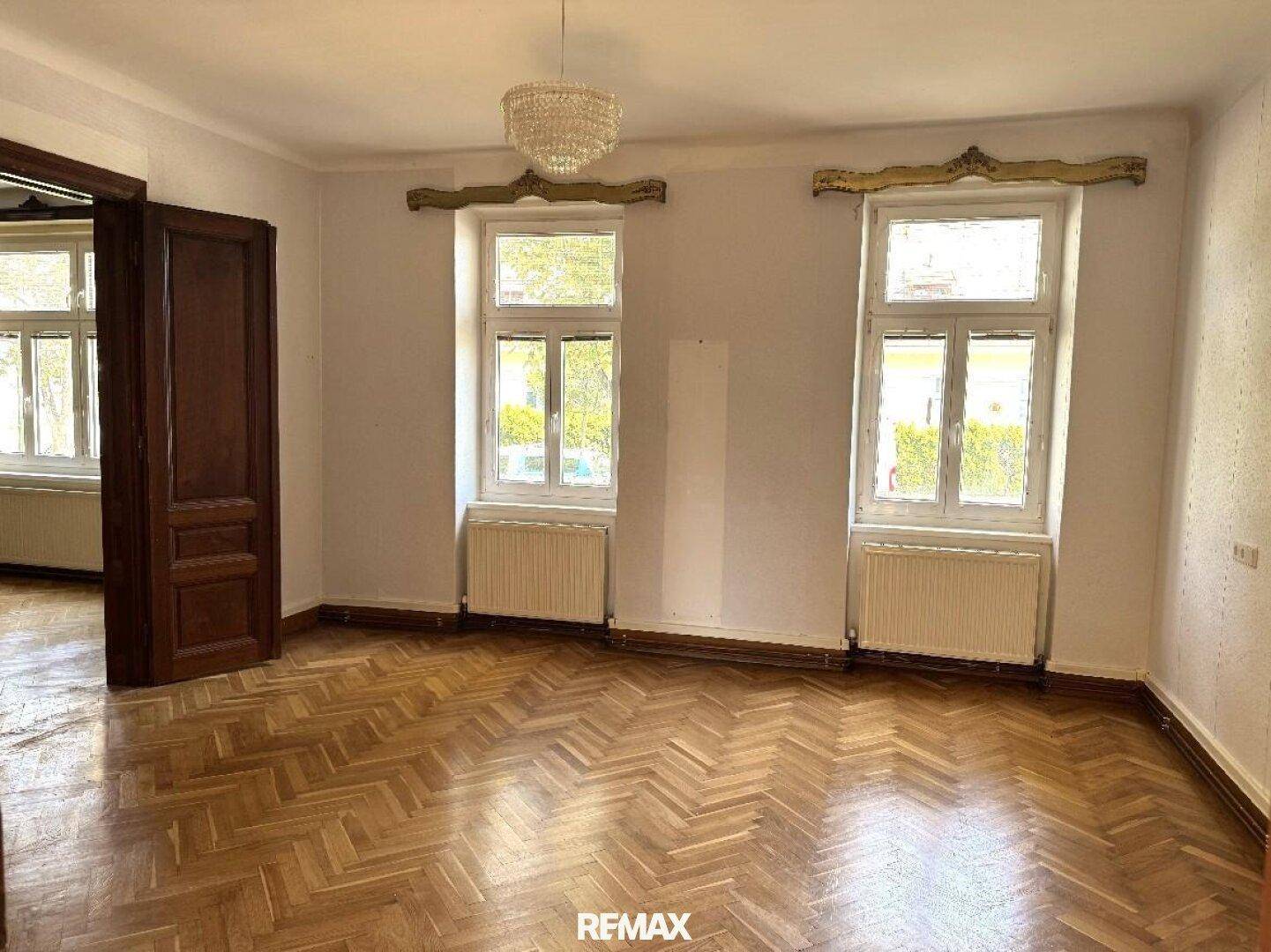 Geräumiges Wohnzimmer mit klassischem Fischgrätparkett, großen Fenstern und einem eleganten Kronleuchter.