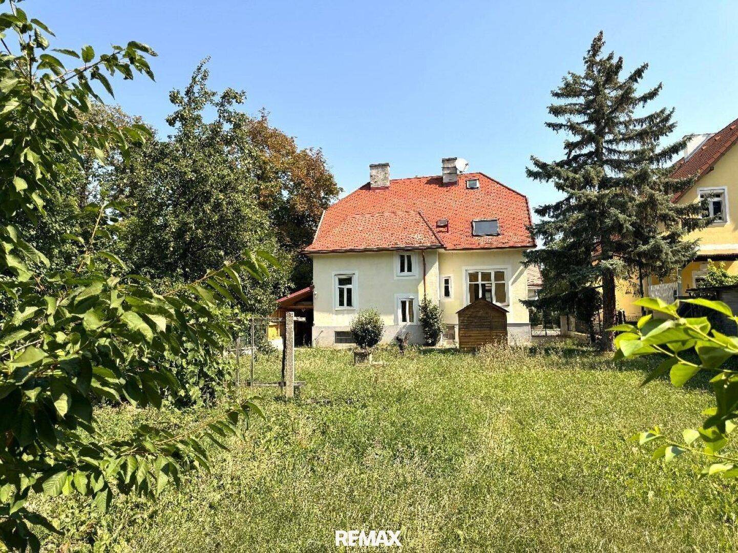 Ansicht des Hauses vom Garten aus, mit gelber Fassade, rotem Dach und üppigem Grün.