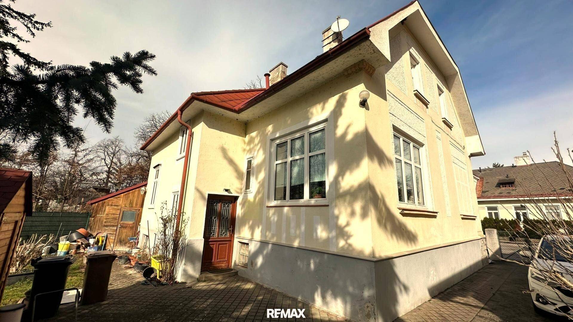 Seitenansicht des Hauses mit Eingangstür, gelber Fassade und gepflasterter Auffahrt unter hellem Himmel.