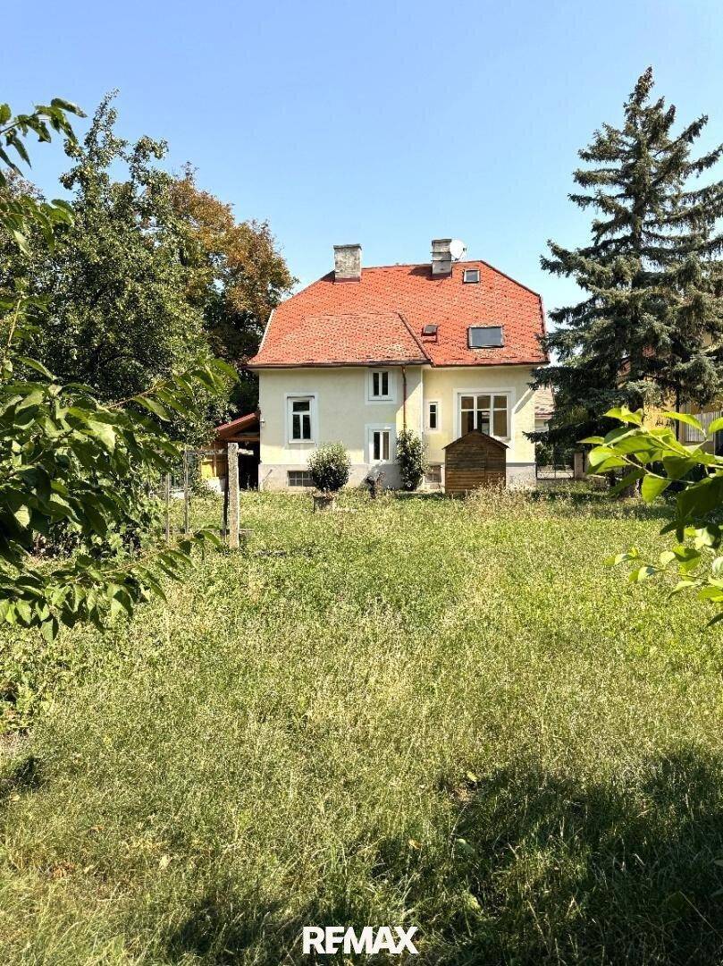 Das Haus von der Gartenseite, umgeben von einem großen Rasen und hohen Bäumen unter blauem Himmel.