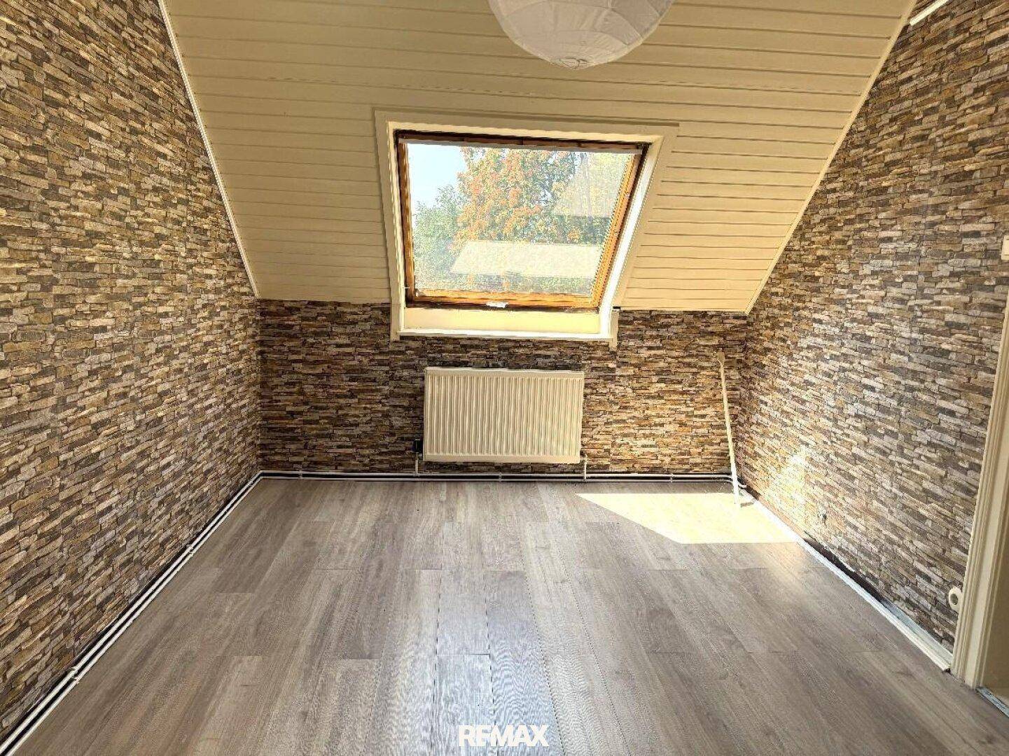 Dachzimmer mit schräger Decke, Wänden in Steinoptik und einem großen Dachfenster für viel Helligkeit.
