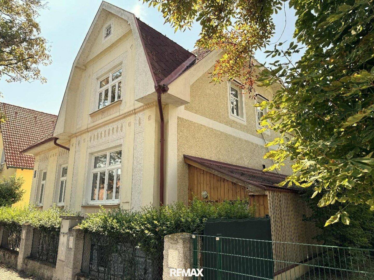 Seitenansicht des Hauses mit gelber Fassade, rotem Dach und einem hölzernen Anbau, umgeben von Bäumen.