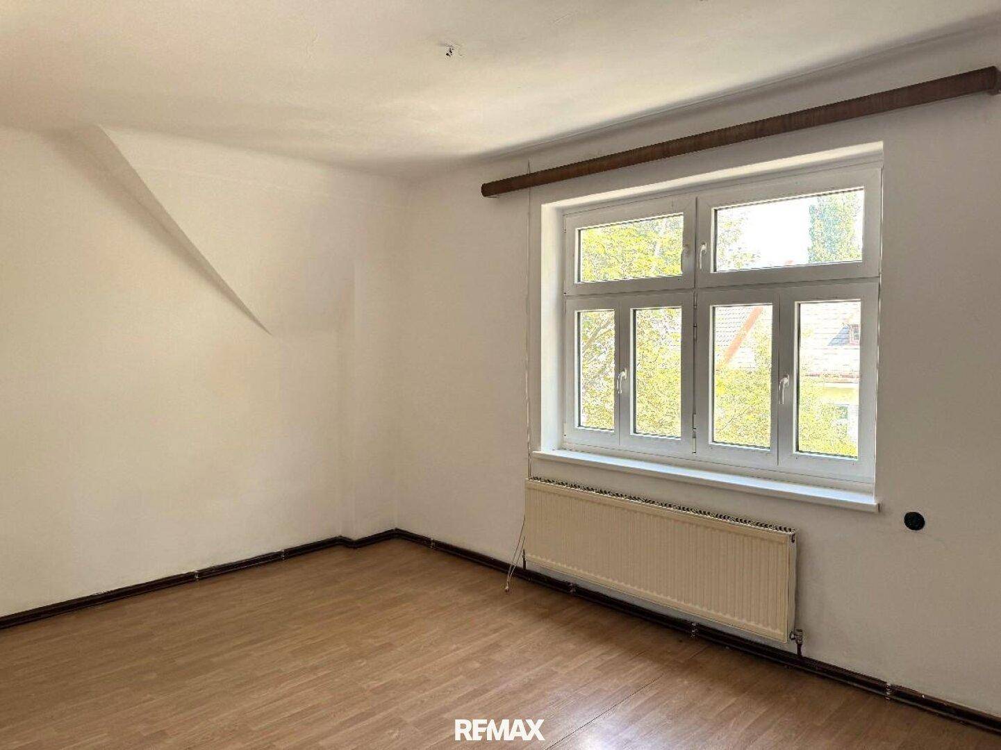 Helles Dachzimmer mit weiß gestrichenen Wänden, großem Fenster und hellem Holzboden, bietet viel Tageslicht.