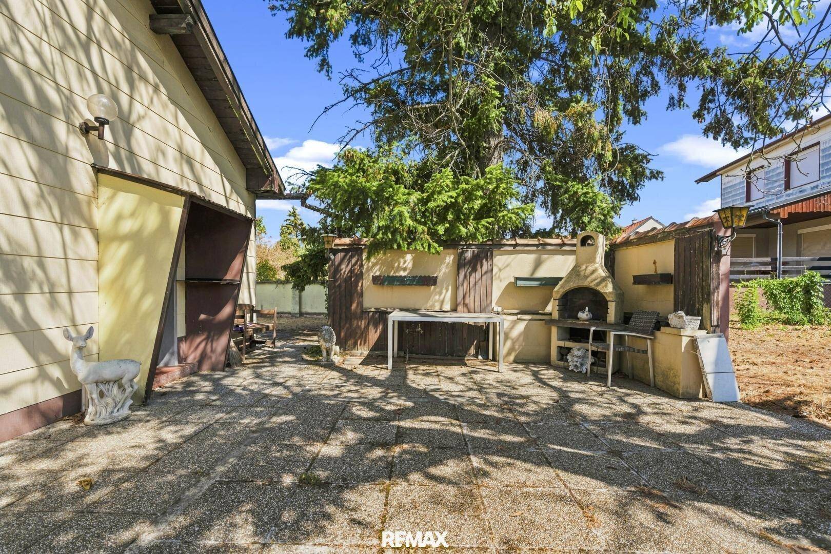 Gepflasterter Terrassenbereich mit gemauertem Grill und Gartenfiguren unter Bäumen.
