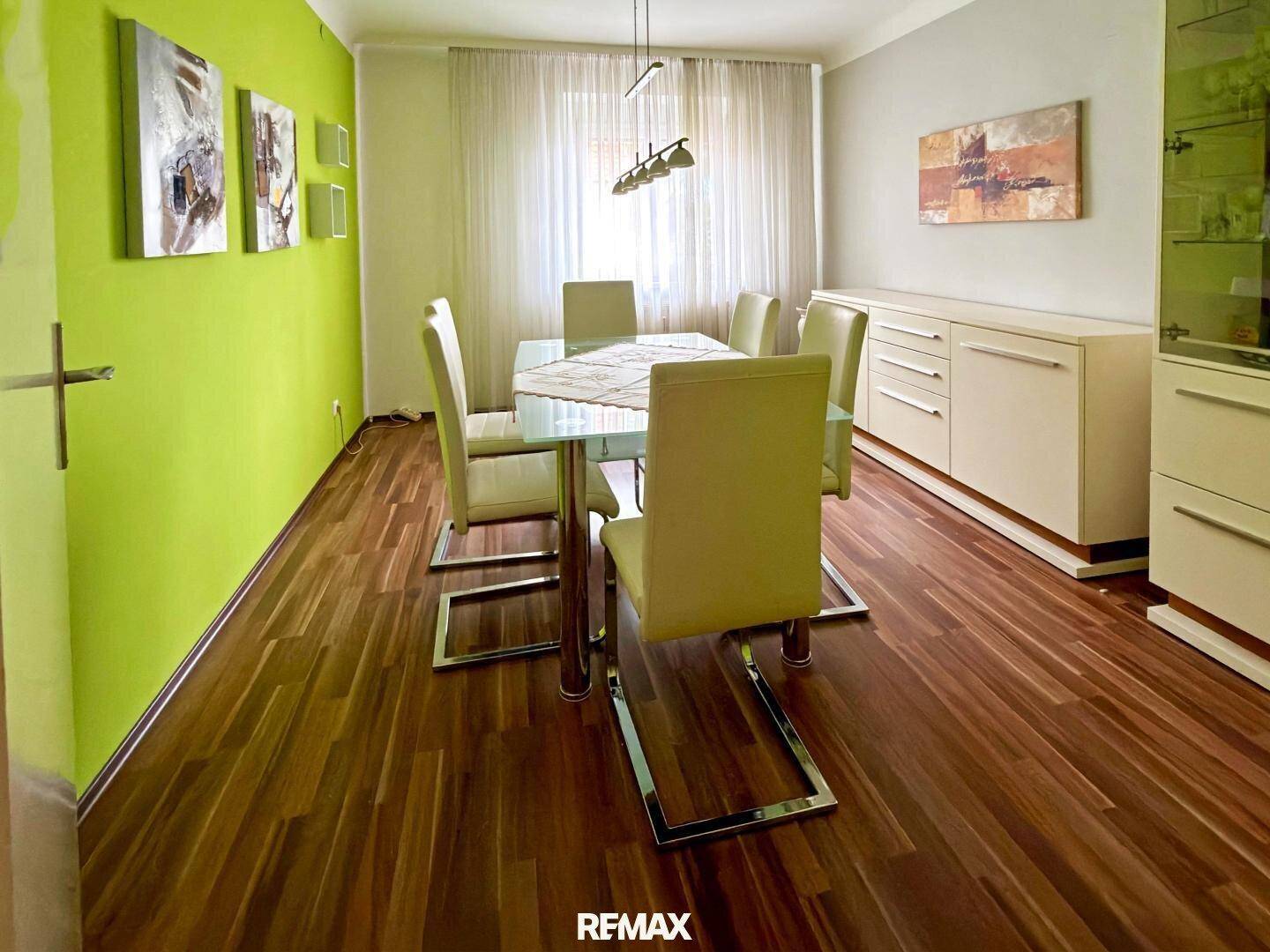 Heller Essbereich mit modernem Glastisch, grüner Akzentwand und Holzfußboden.