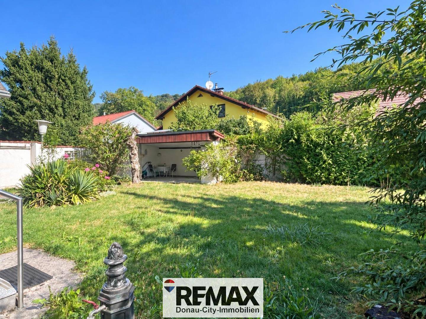 Großer, sonniger Gartenbereich mit Rasenfläche und überdachtem Stellplatz im Hintergrund.