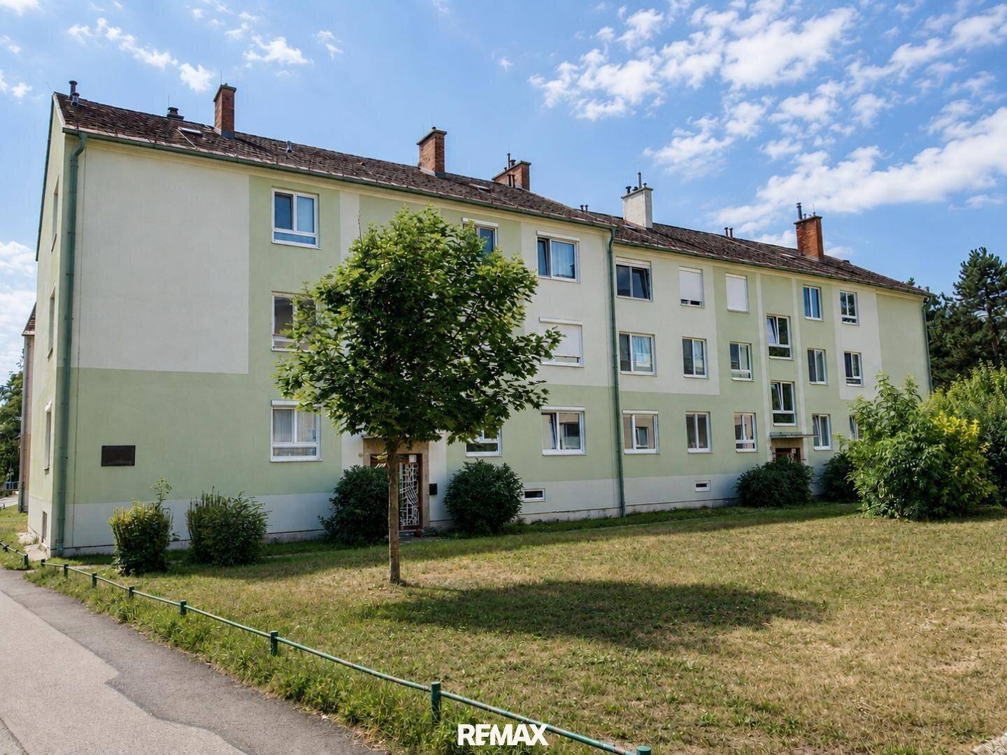 Mehrfamilienhaus mit grüner Fassade, vielen Fenstern und umliegender gepflegter Grünfläche.