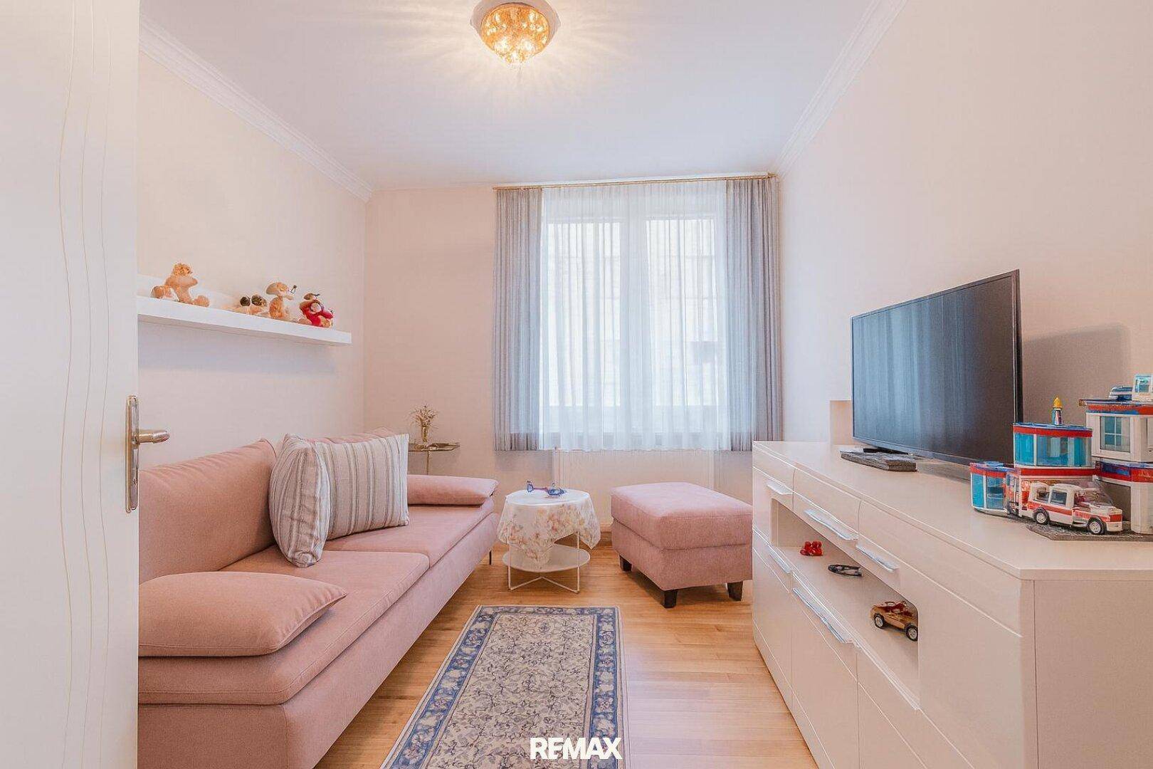Helles Zimmer mit rosa Sofa, Holzfußboden und Spielzeugdekoration, ideal als Kinder- oder Gästezimmer.