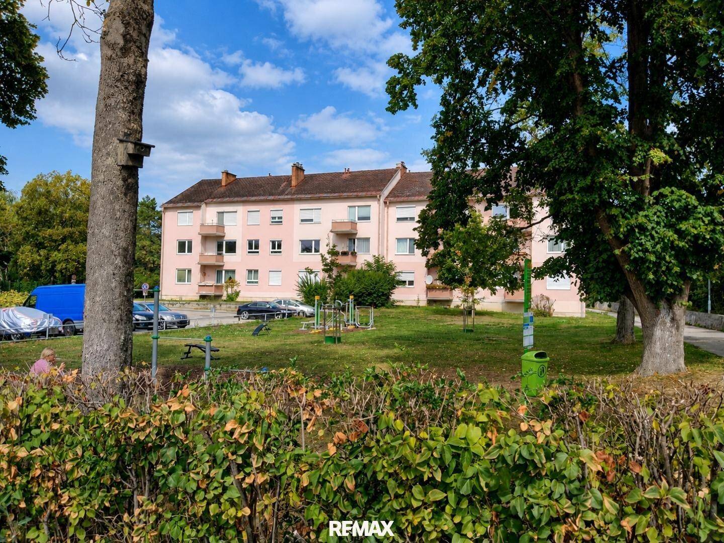 Wohngebäude mit rosa Fassade, Balkonen und einem Spielplatz in der Nähe, umgeben von Bäumen.