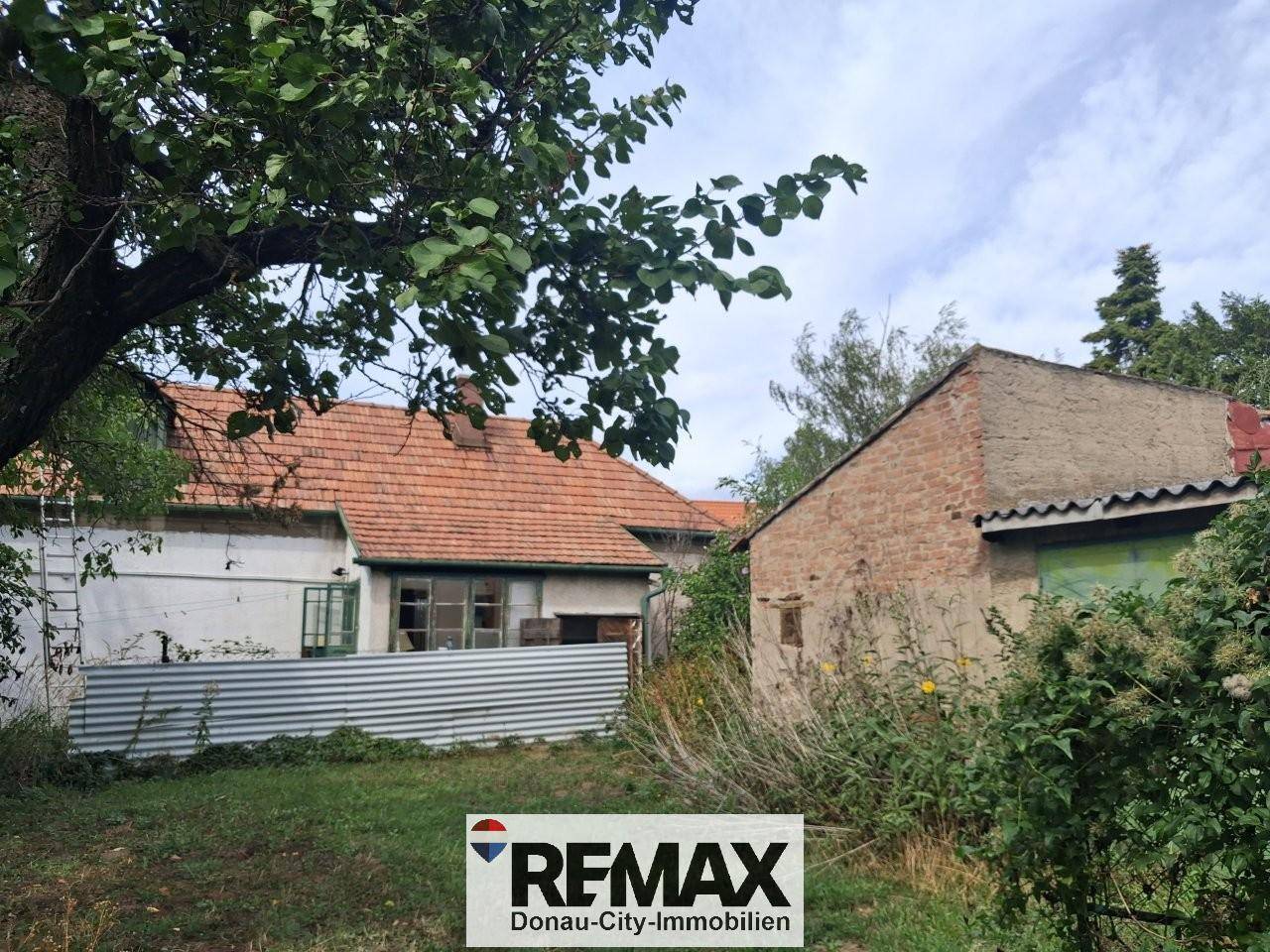 Rückansicht eines alten Hauses mit Ziegeldach und einem Nebengebäude, umgeben von Vegetation.