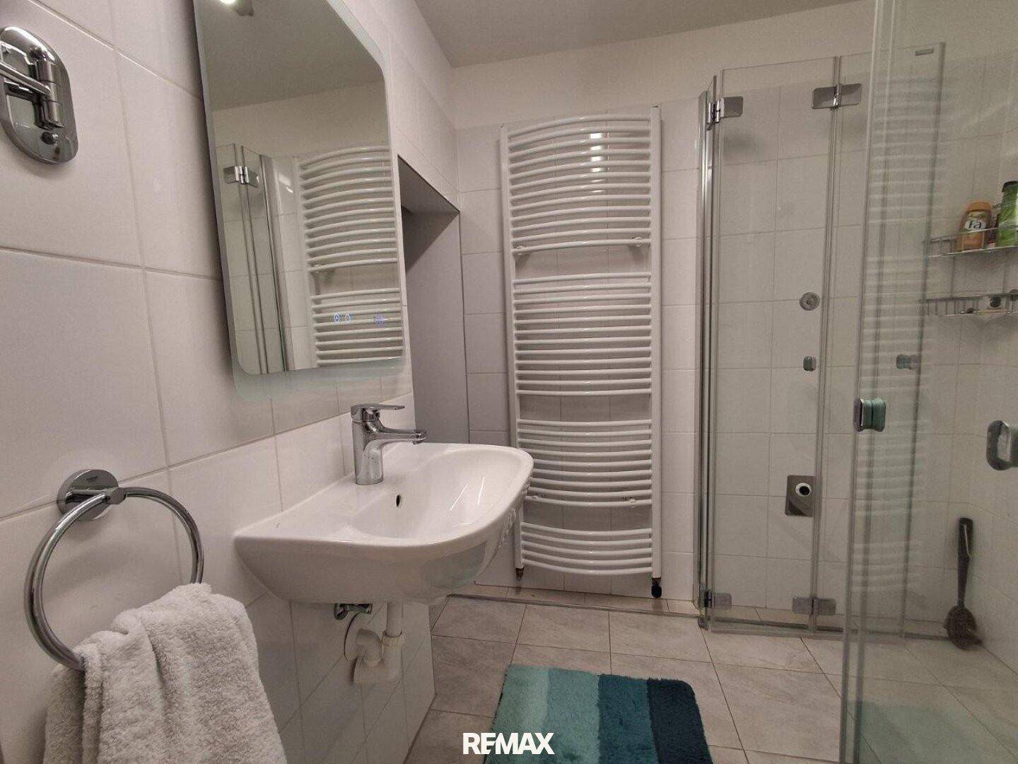 Modernes Badezimmer mit Dusche, Waschbecken, beheiztem Handtuchhalter und hellen Fliesen.