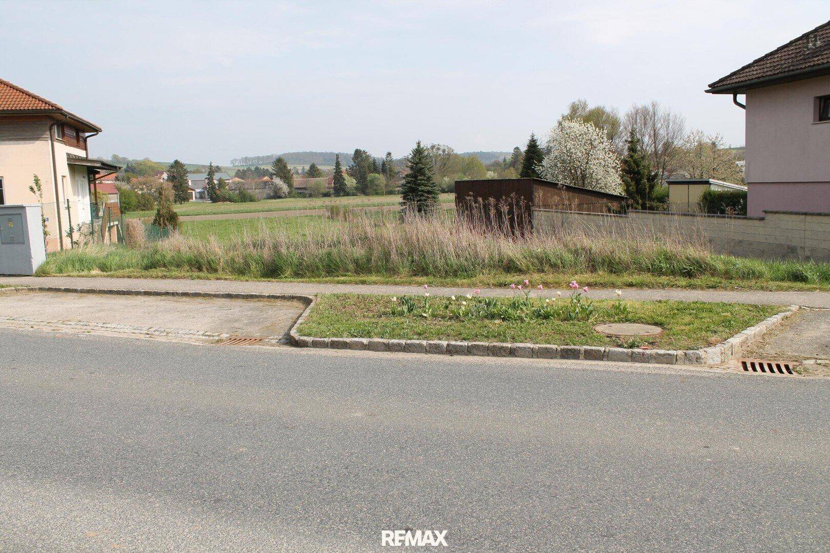 Blick von der Straße auf ein unbebautes Grundstück mit Gehweg und angrenzenden Häusern in ländlicher Umgebung.