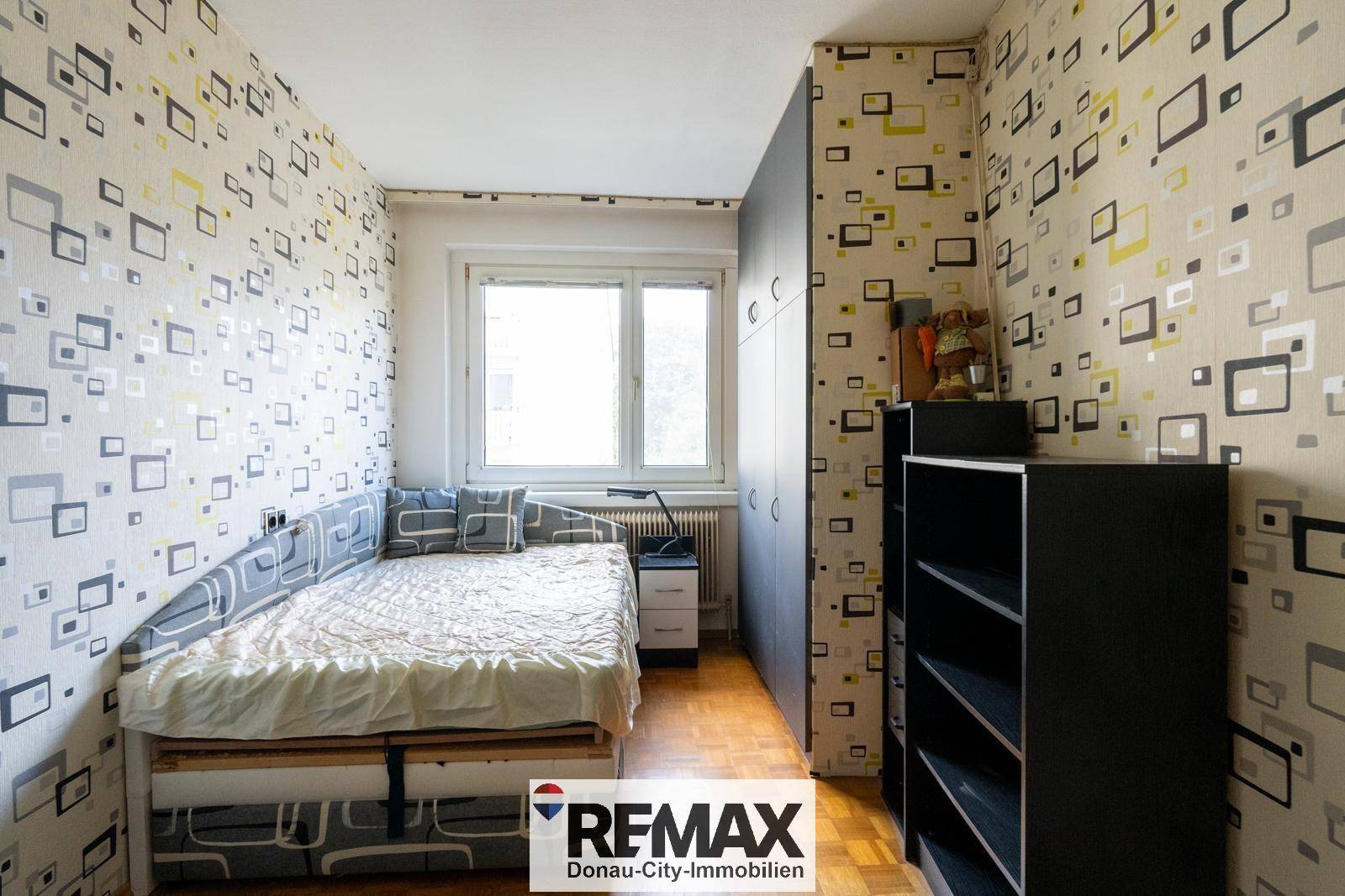 Schlafzimmer mit Einzelbett, Fenster und gemusterter Tapete, ausgestattet mit Parkettboden.