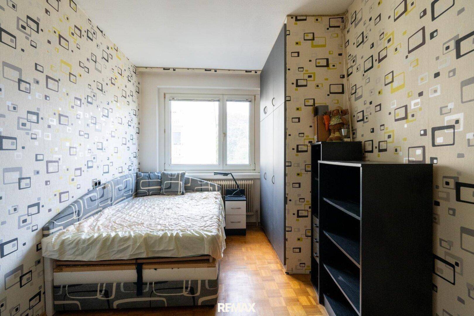Schlafzimmer mit Einzelbett, Parkettboden und gemusterter Tapete.