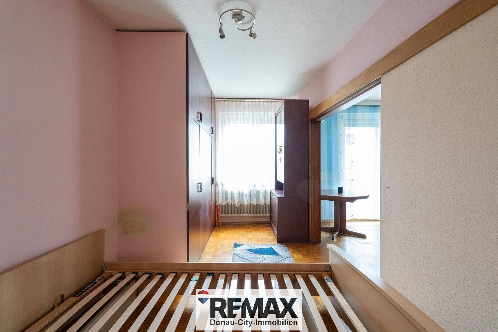 Schlafzimmer mit Bettrahmen, rosa Wänden und Kleiderschrank, angrenzend an einen weiteren Raum.