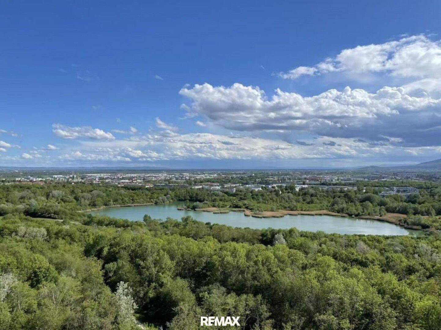 Malerischer Weitblick über einen See und die umliegende grüne Landschaft unter blauem Himmel.