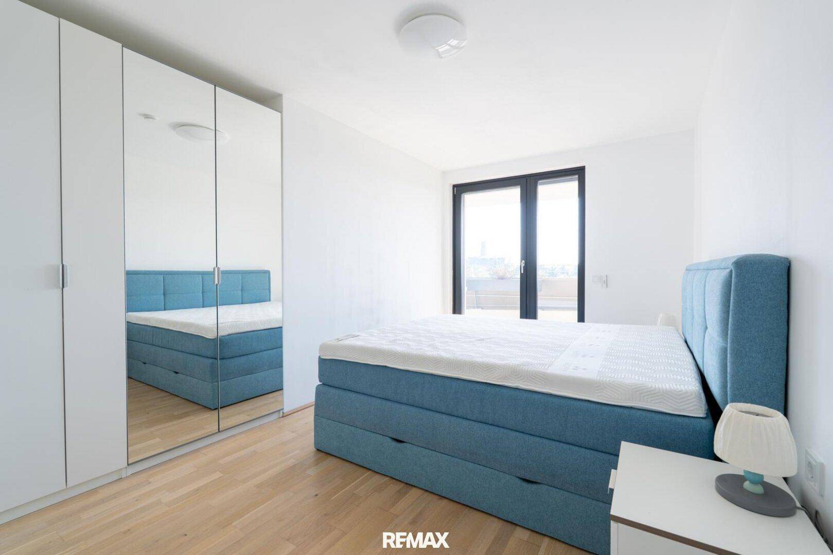 Helles Schlafzimmer mit blauem Boxspringbett, Spiegelschrank und Holzboden.