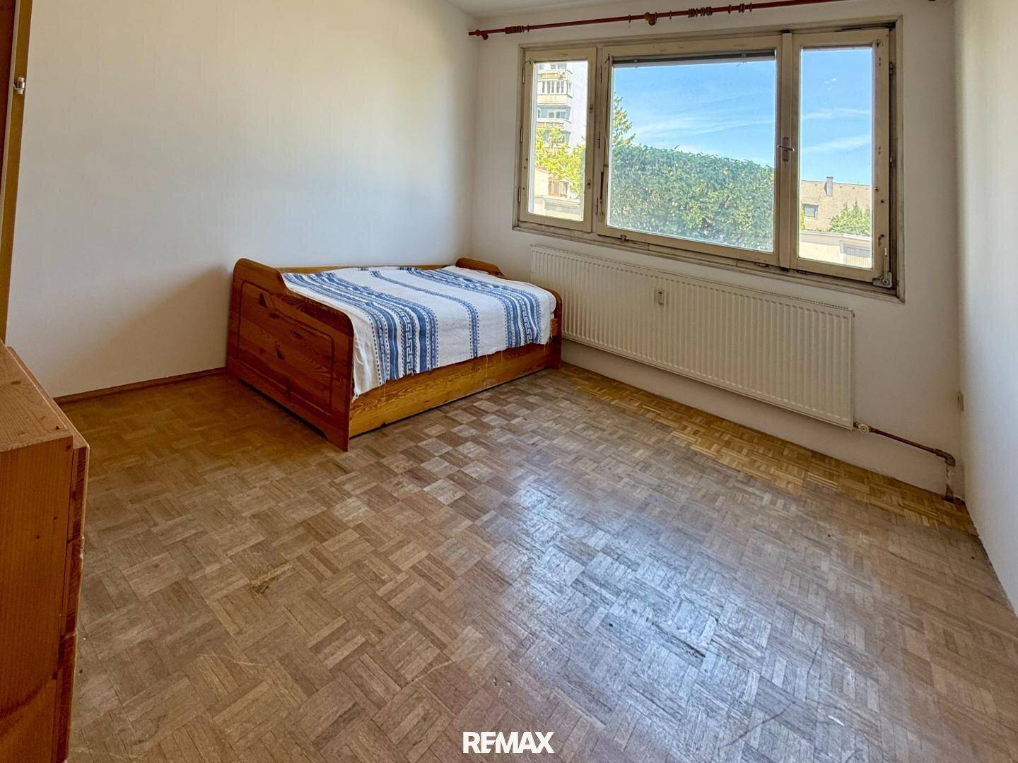 Einfaches Schlafzimmer mit Holzbett, Parkettboden und Fenster mit Stadtblick.
