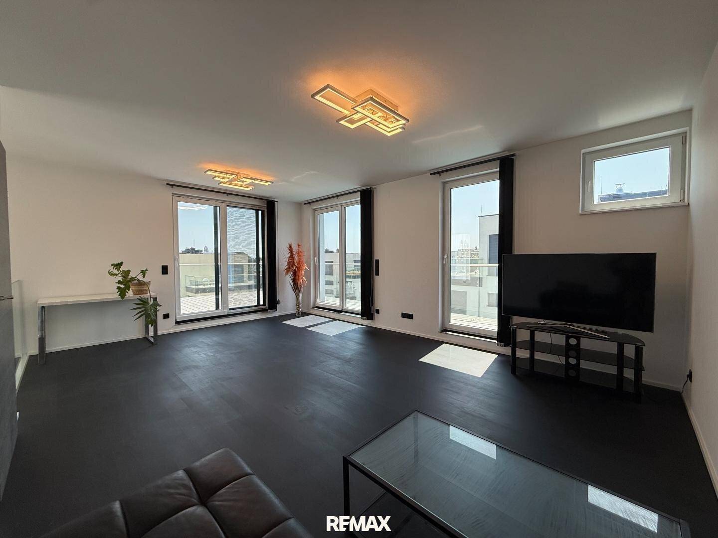 Helles Wohnzimmer mit dunklem Boden, großen Fenstern und Zugang zu einem Balkon.