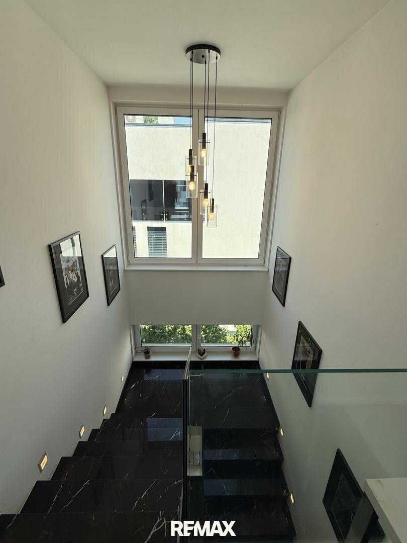 Hohes Treppenhaus mit moderner Hängelampe, großen Fenstern und viel Tageslicht.