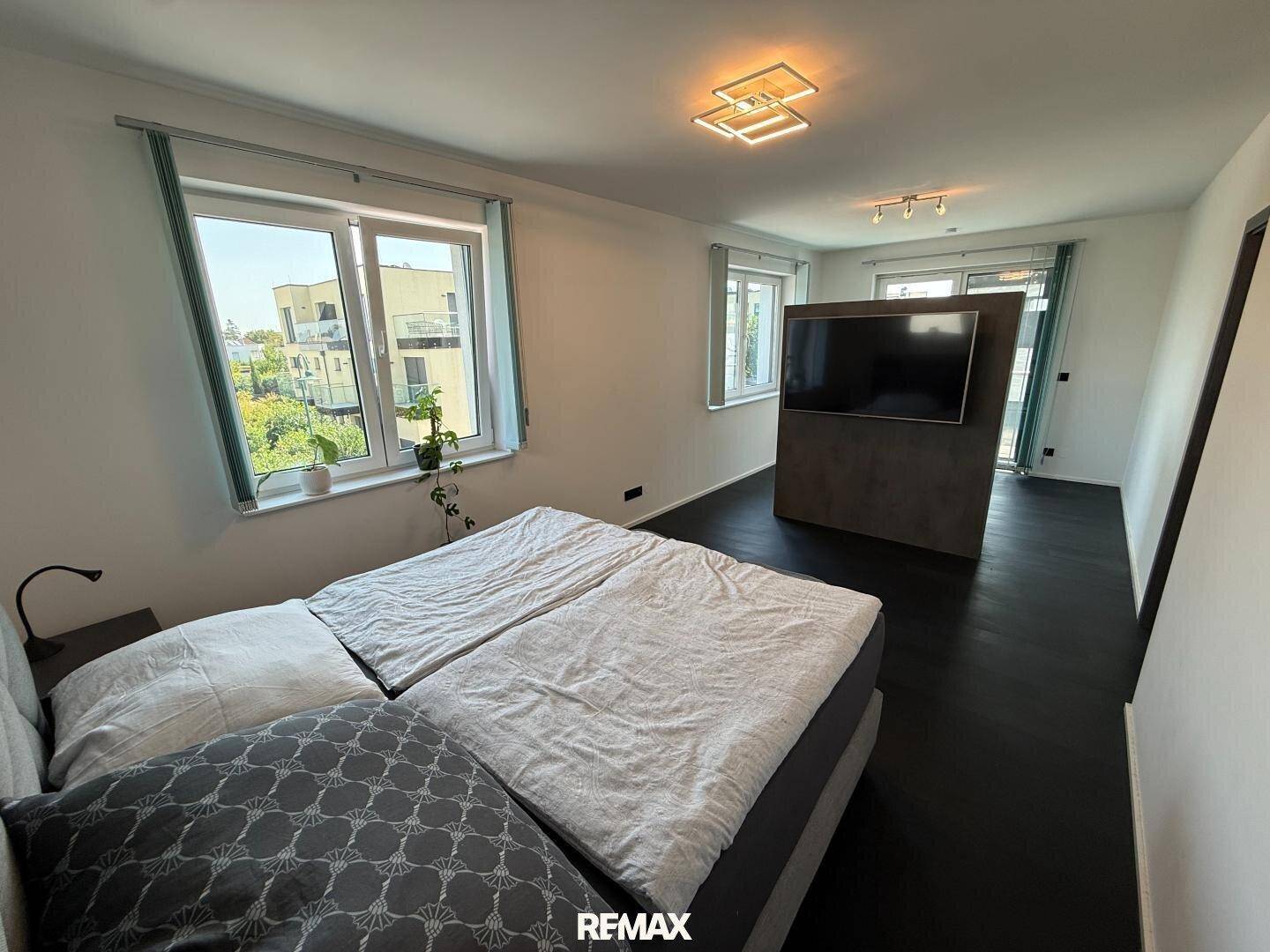 Helles Schlafzimmer mit großem Bett, dunklem Boden und Fenstern mit Blick ins Grüne.