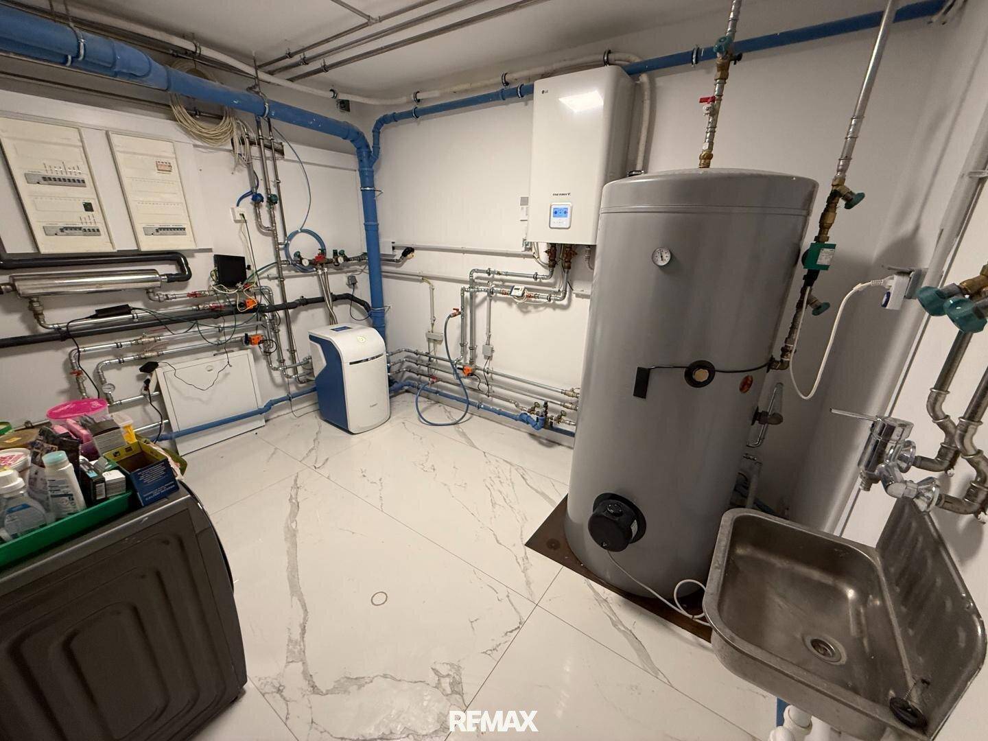 Technikraum mit Warmwasserspeicher, umfangreicher Verrohrung und Waschbecken.