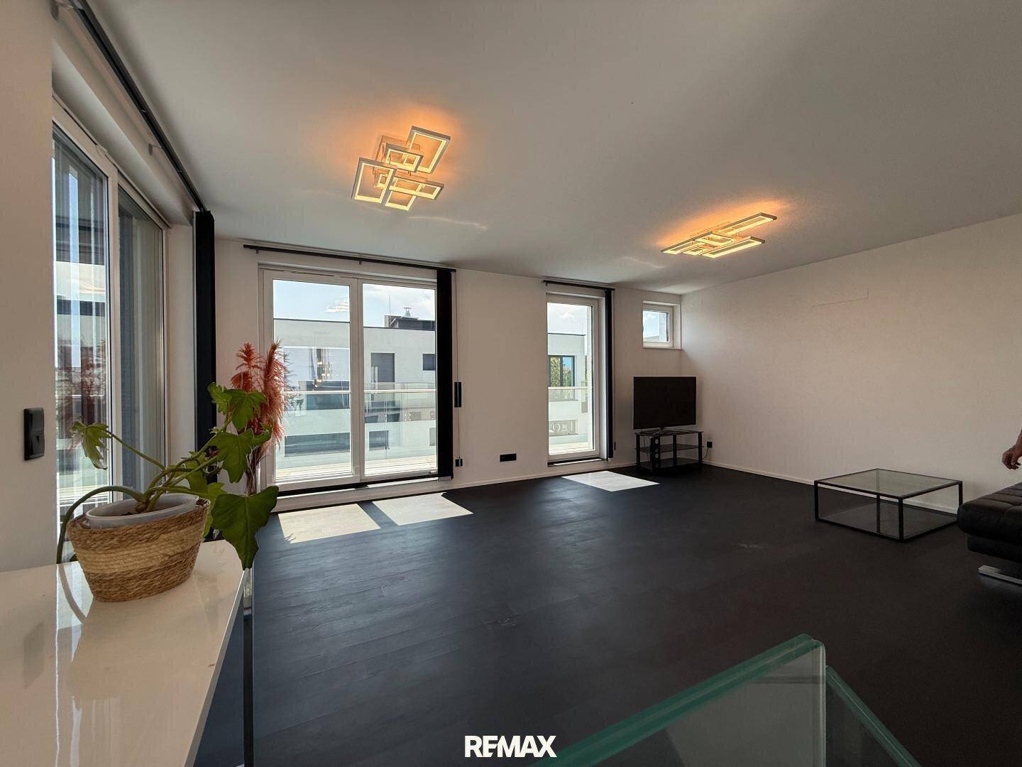 Geräumiges Wohnzimmer mit dunklem Boden, großen Fenstern und viel Tageslicht.
