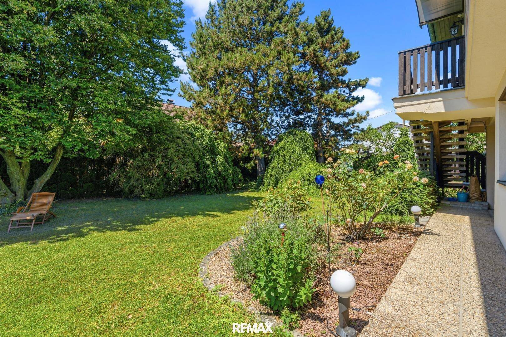 Gartenweg mit Blumenbeeten, Rasenfläche und hohen Bäumen.