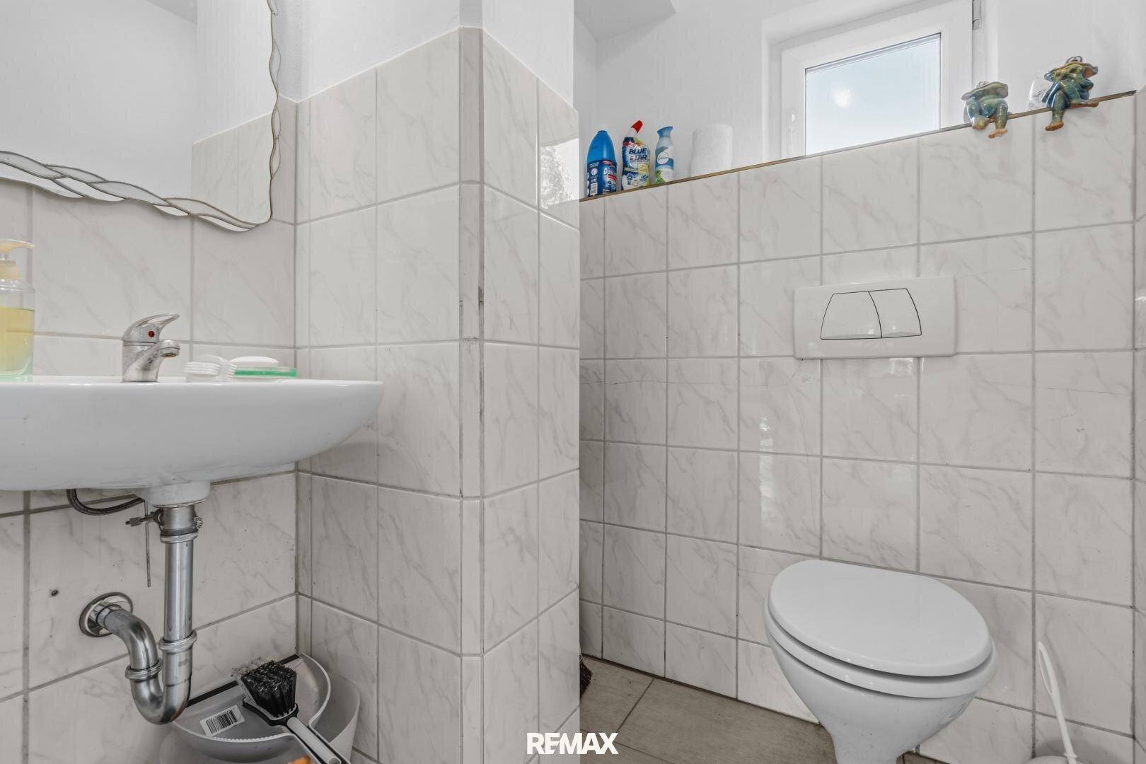 Kleines WC mit weißen Fliesen, Waschbecken und Fenster für Tageslicht.