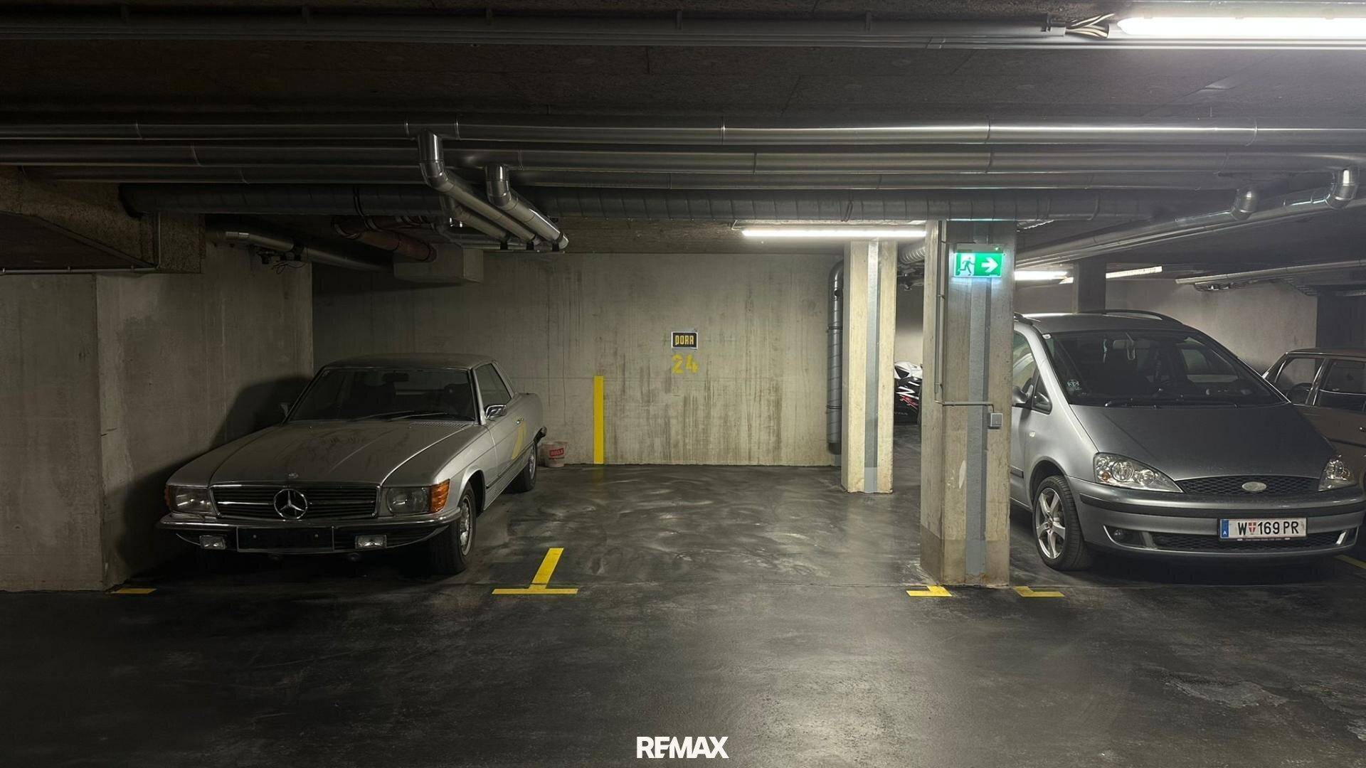 Tiefgarage mit zwei geparkten Autos, Betonwänden und sichtbaren Versorgungsleitungen an der Decke.