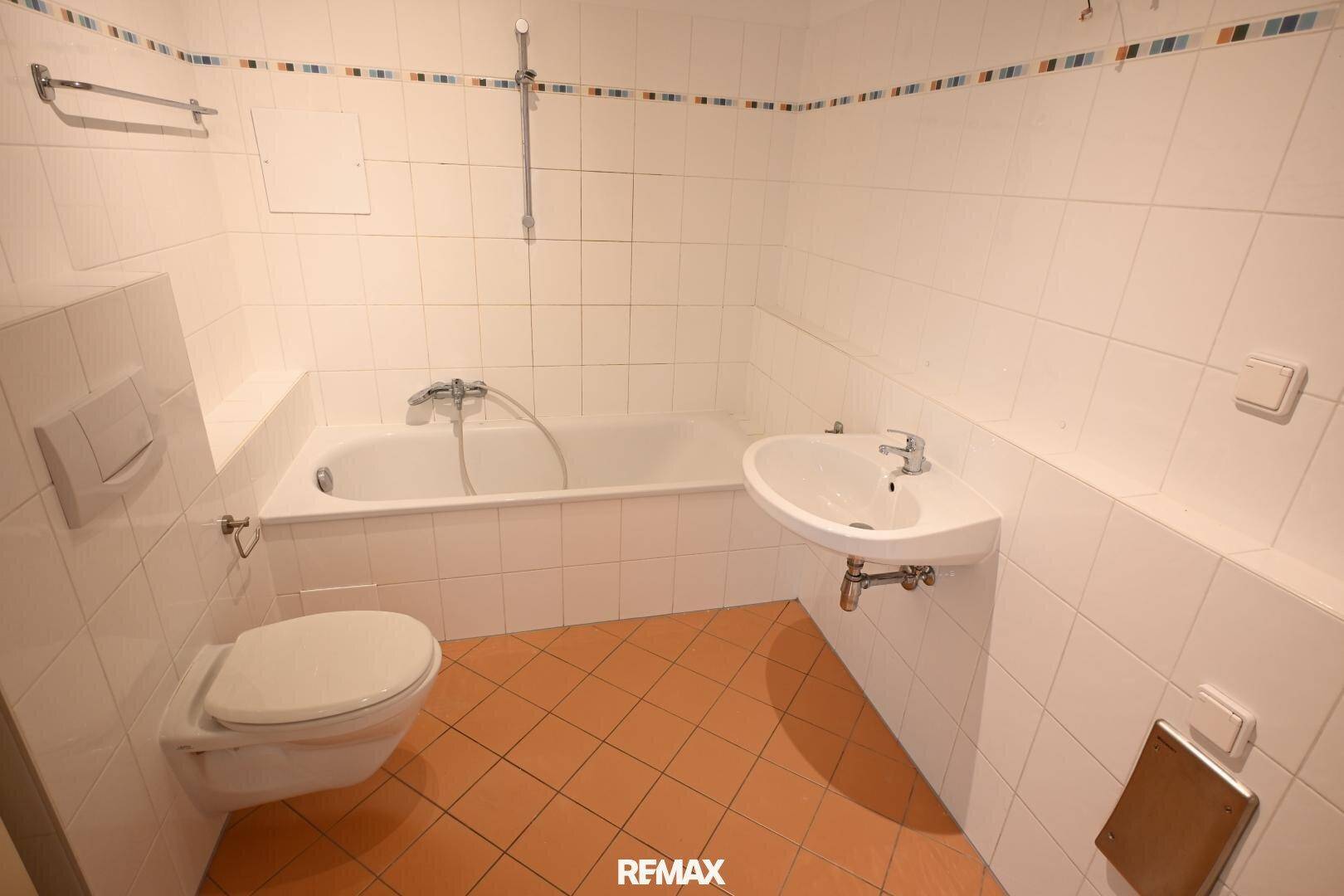 Badezimmer mit Badewanne, Toilette, Waschbecken und weißen Fliesen sowie Terrakotta-Bodenfliesen.