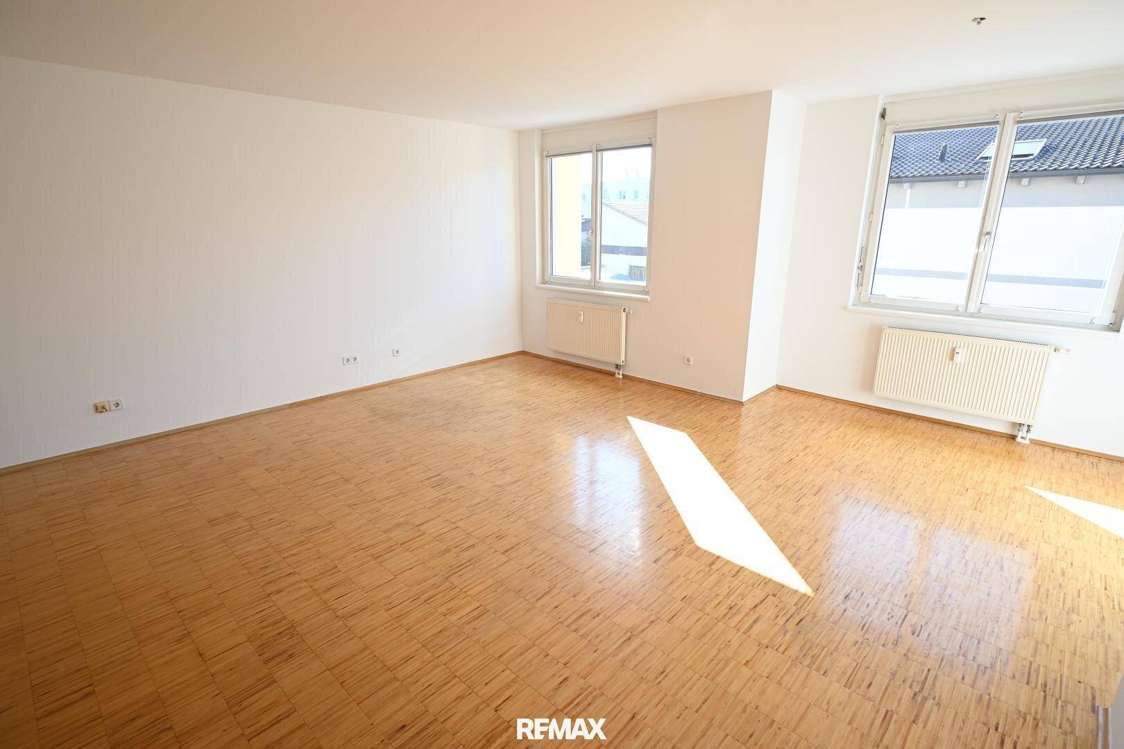Heller, leerer Wohnbereich mit Holzfußboden und großen Fenstern, die den Raum mit Sonnenlicht füllen.