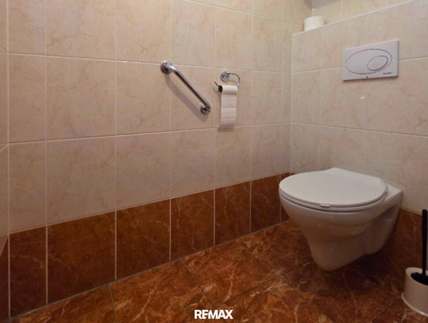 Separate Toilette mit Wand-WC, Haltegriff und beigefarbenen Fliesen.