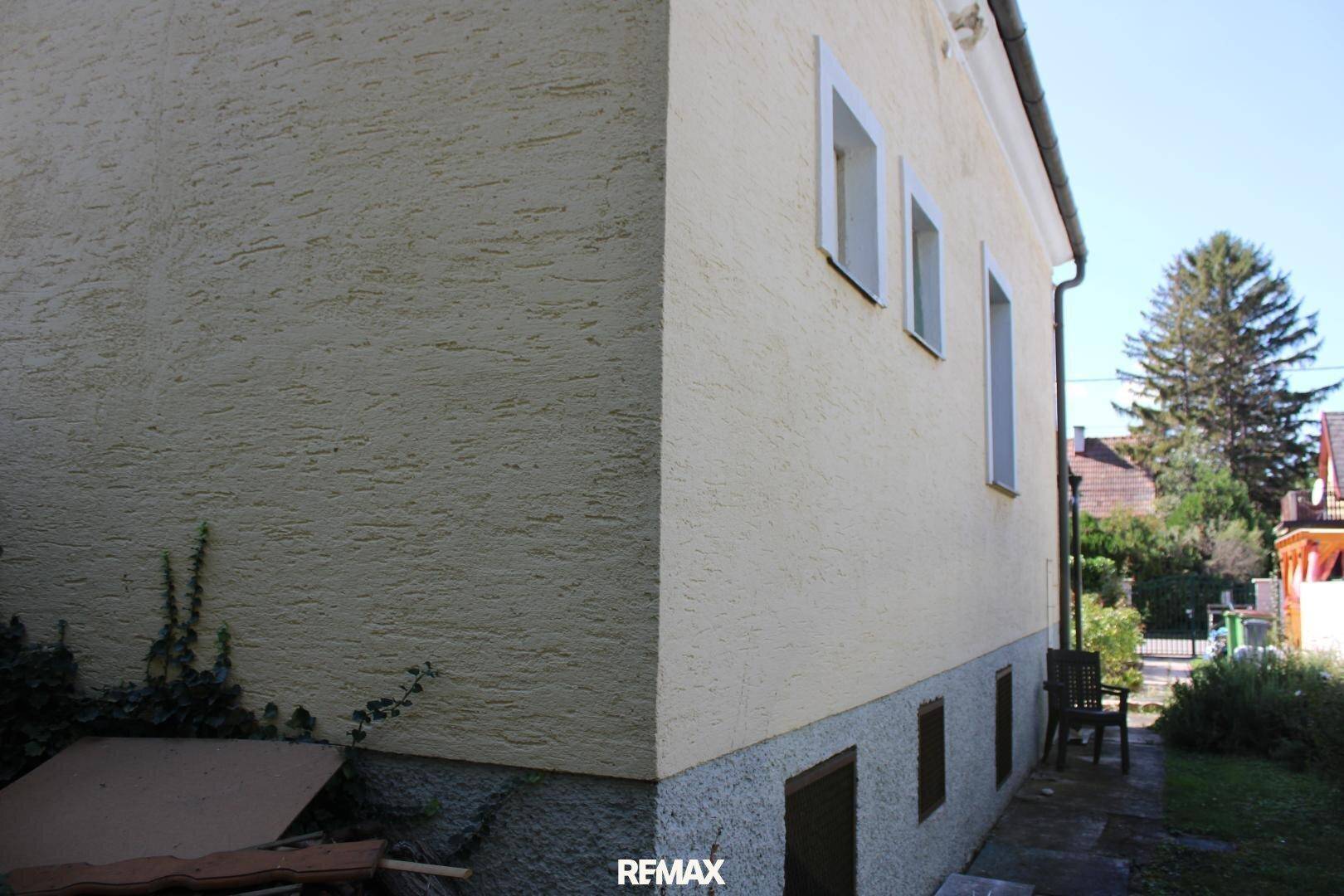 Seitenansicht des Hauses mit rauer Putzfassade und kleinen Fenstern, umgeben von Grün.