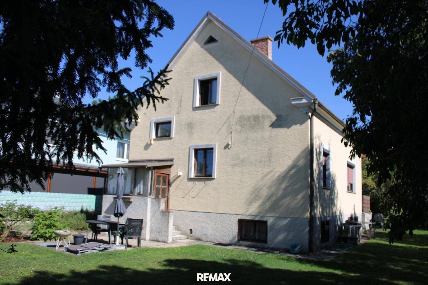 Rückseite des Einfamilienhauses mit Garten, Terrasse und umgebenden Bäumen unter blauem Himmel.