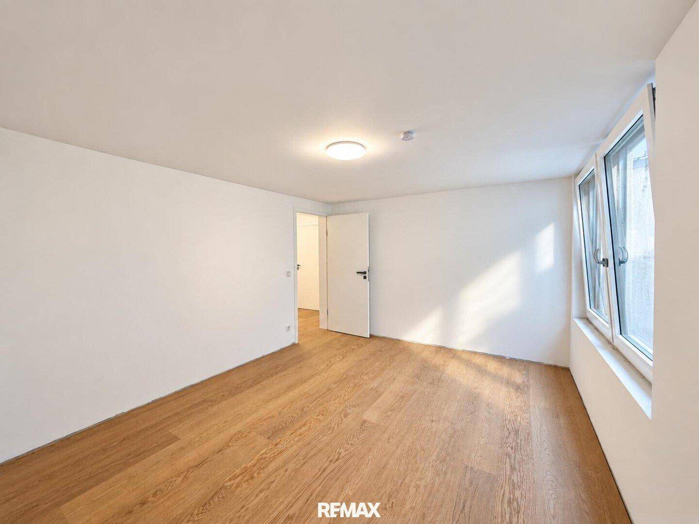 Helles, leeres Zimmer mit Holzboden und großem Fenster, vielseitig nutzbar.